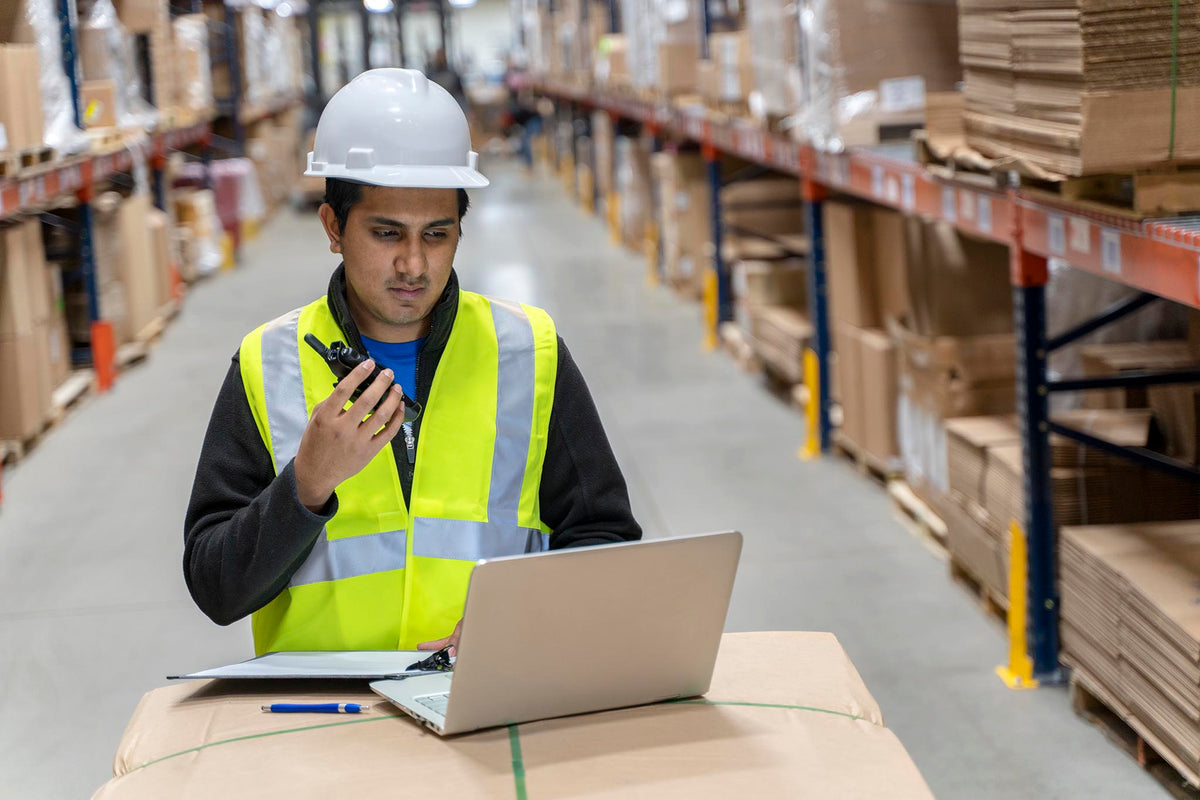 Warehouse employee in a hard hat and high-visibility vest holding a radio beside a laptop and clipboard.