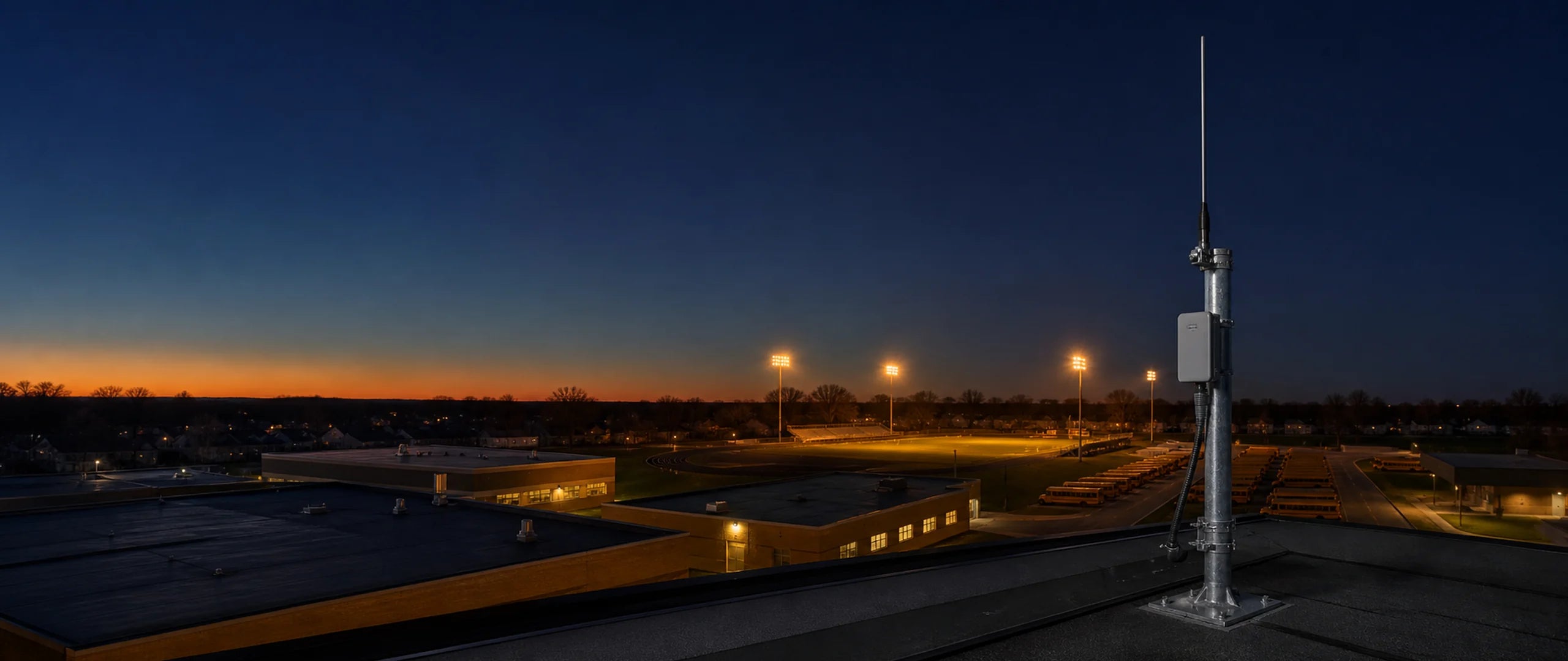 School campus at dusk with rooftop antenna infrastructure, illuminated athletic field, and parked school buses